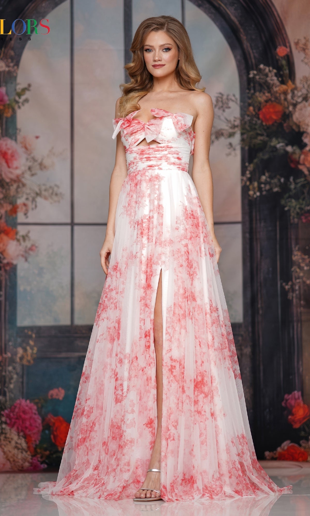 Woman in a red Colors Dress 3811 long prom dress with strapless neckline, a-line skirt, and front slit stands indoors before an arched window and floral backdrop.